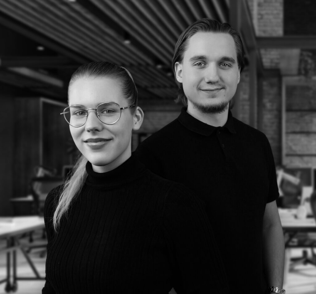 Ein Mann und eine Frau die eine Brille auf hat stehen in einem Büro und lächeln in die Kamera